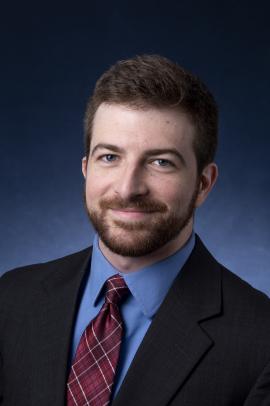 Headshot of Andrew Gray wearing a black blazer, blue button down, and plaid burgundy necktie against a navy blue ombre background